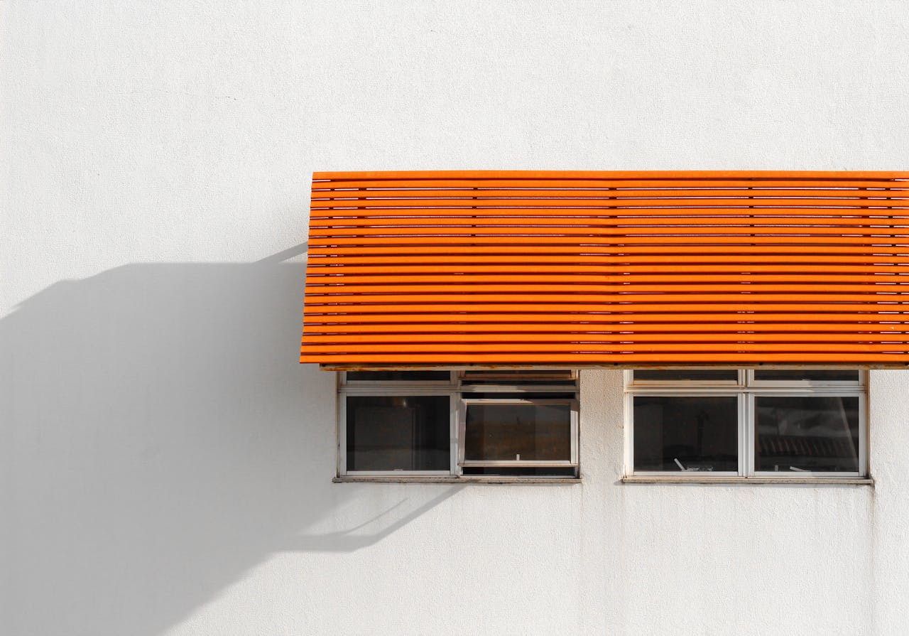 A modern orange awning casts shadows on a white building facade with windows.